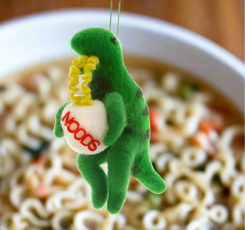 The Canton Christmas Shop Cody Foster Dinosaur with Ramen Noodles Soup Ornament in front of bowl of ramen