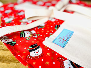 The Canton Christmas Shop Frost the Snowman Christmas Baking Cooking Apron Red Background Gold Snowflakes Corn Cob Pipe Button Nose Two Eyes Made Out of Coal by Two Medium Sized Ladies close up view